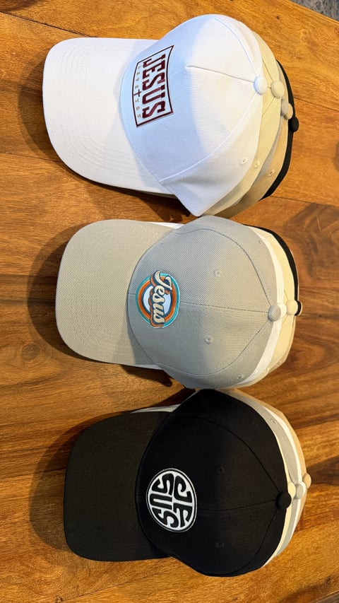 Three baseball caps stacked vertically on a wooden surface: white with red logo on top, light gray with blue Bud Light logo in middle, black with white circular logo on bottom