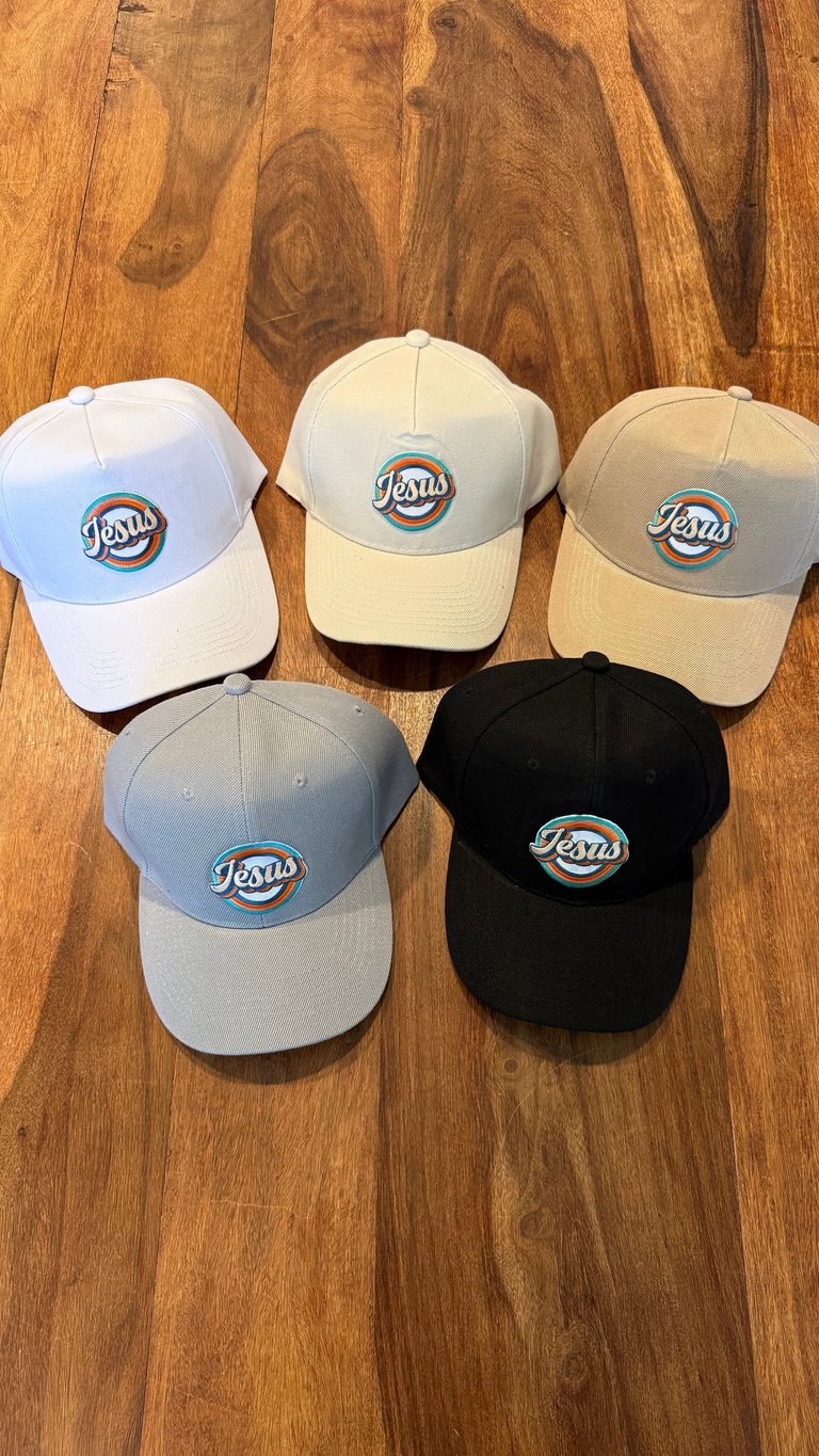 Five Jesus baseball caps in different colors arranged on a wooden surface