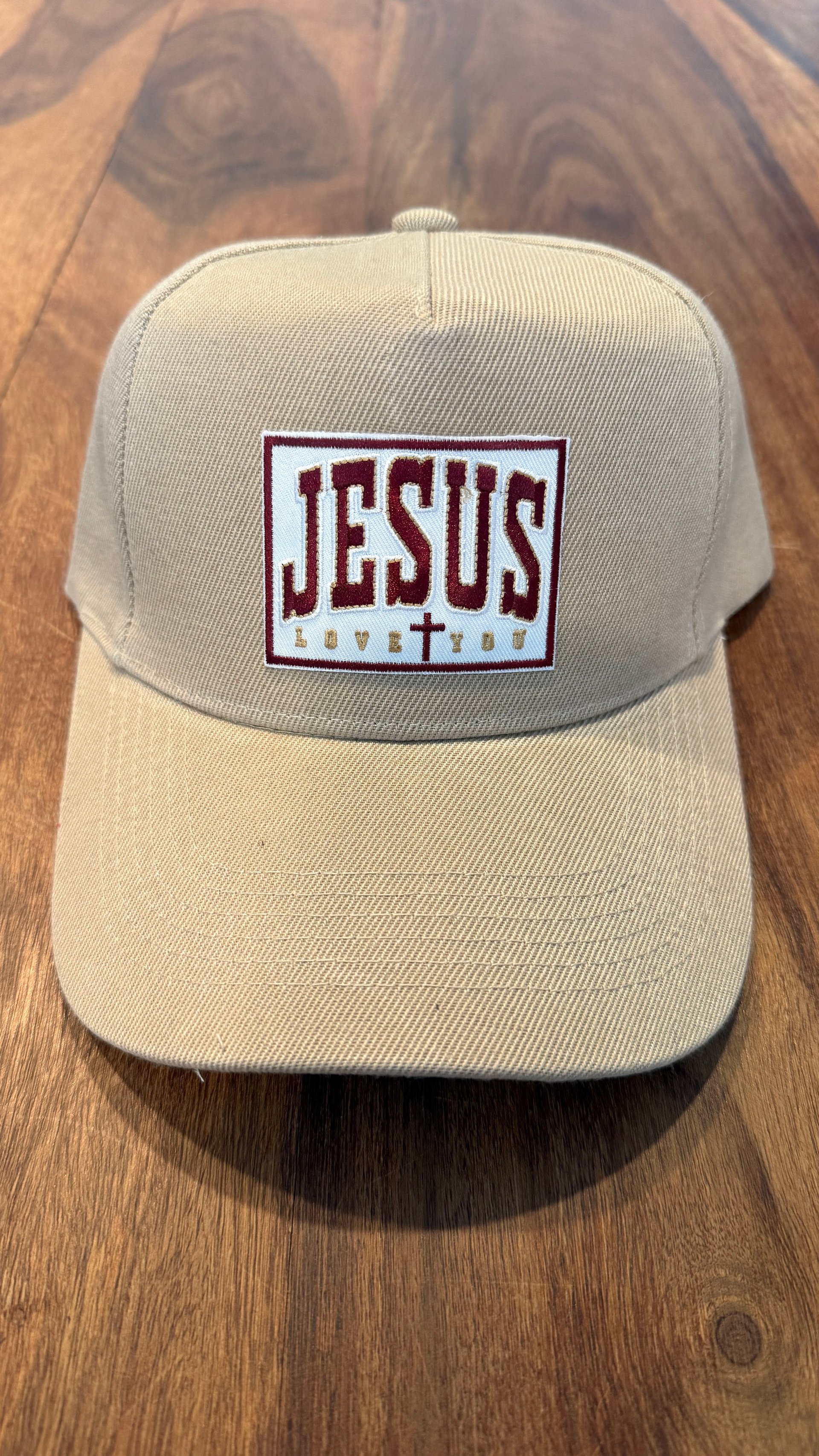 Beige baseball cap with maroon Jesus Saves text and cross symbol on patch, placed on wooden surface