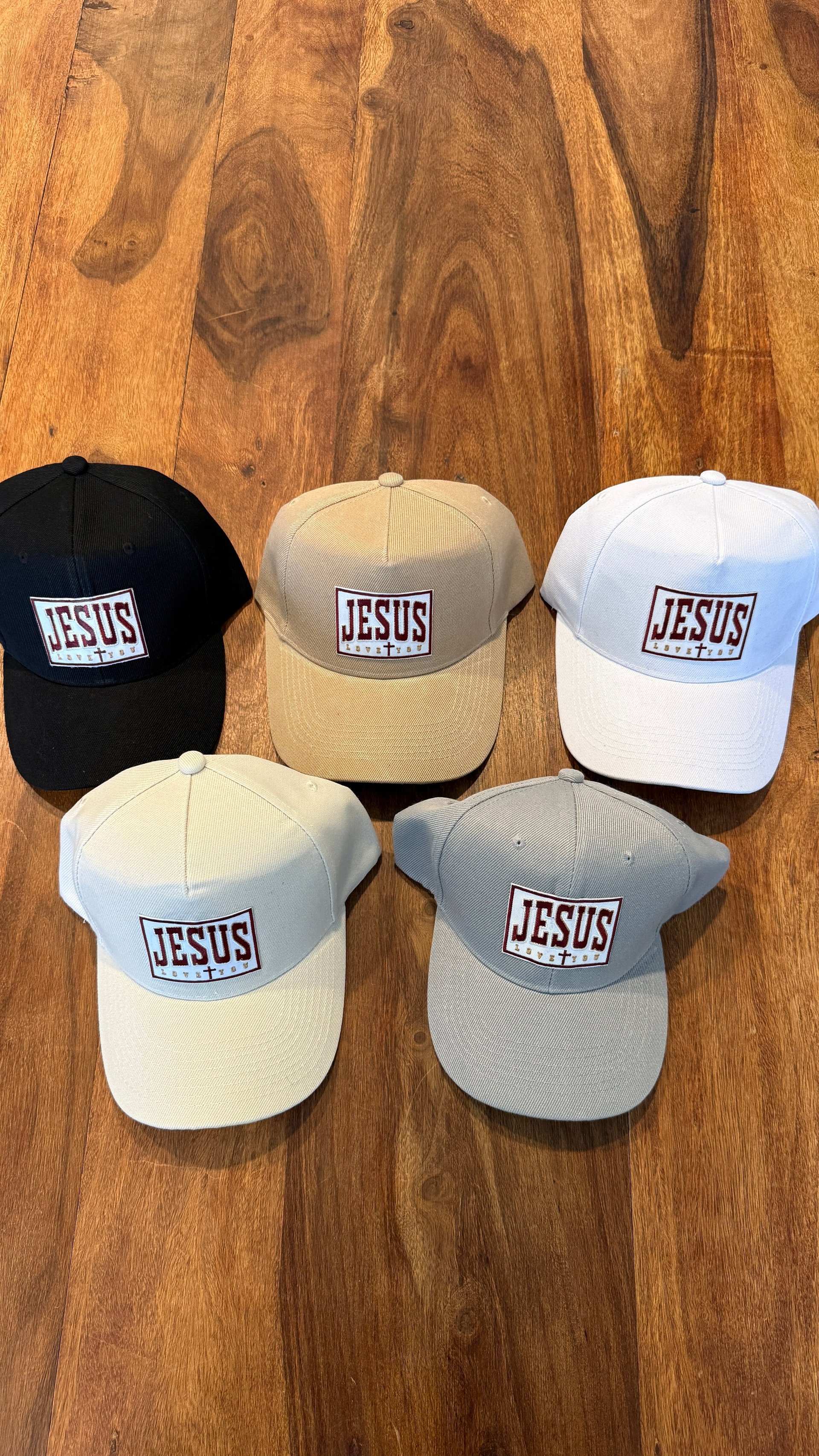 Five JESUS baseball caps in black, beige, white, cream, and gray arranged on a wooden floor